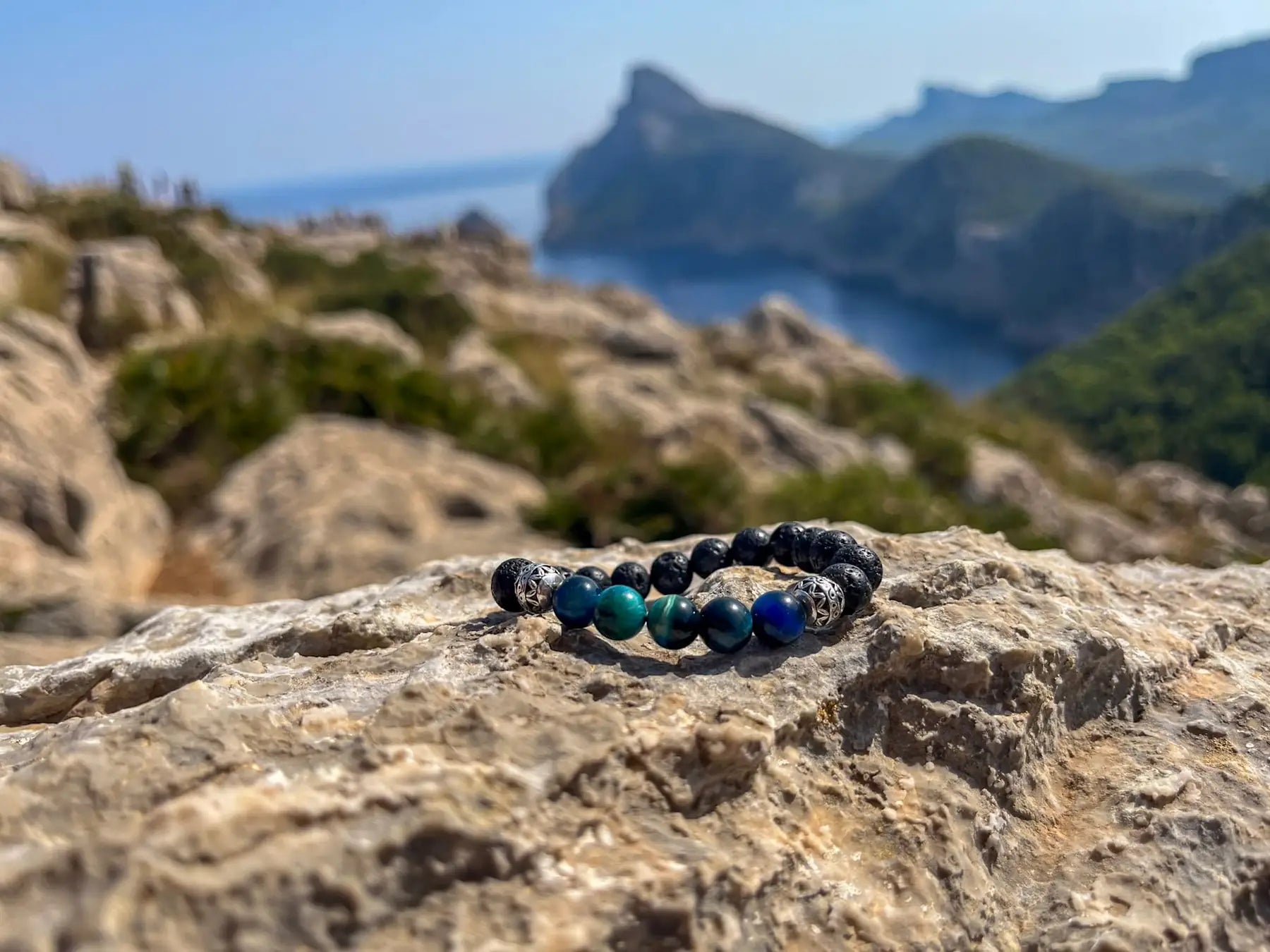Armband aus Natursteinen auf Felsen mit Meereslandschaft im Hintergrund.
